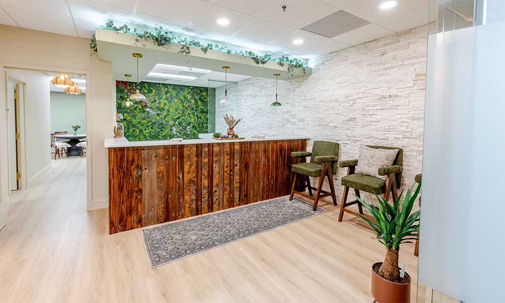 Reception area with wooden counter, green chairs, plant, and decorative foliage on the wall.