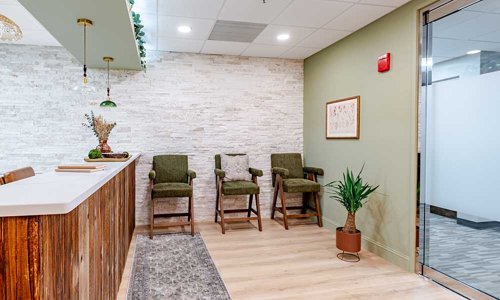 Reception area with stone wall, wooden counter, hanging lights, green chairs, framed artwork, and potted plant.