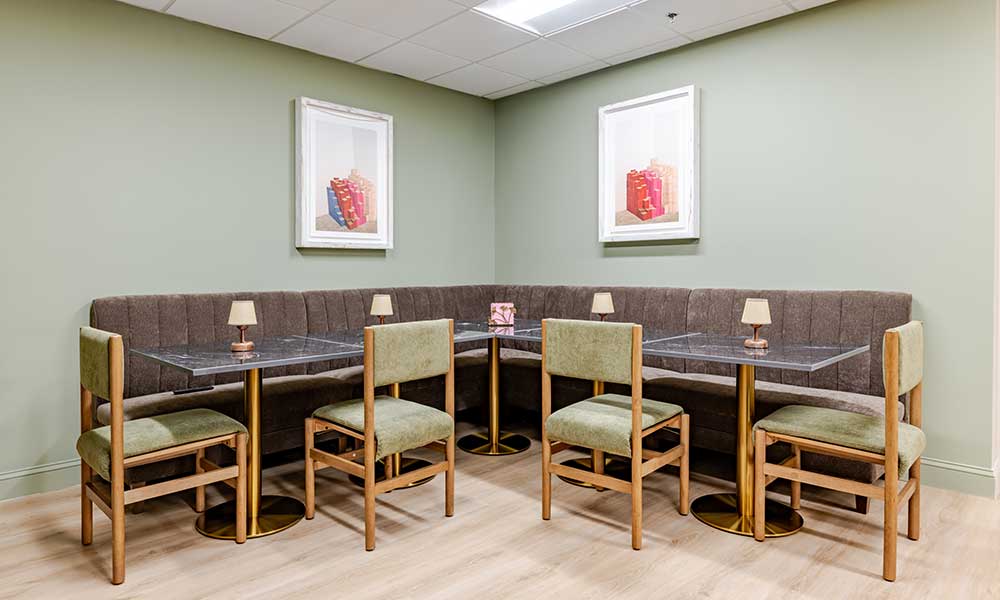 Dining area with green chairs, glass tables, brown bench, and framed artwork on pale green walls.