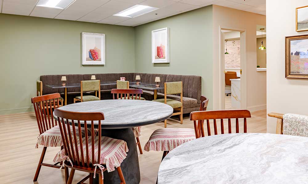 Dining area with round marble tables, wooden chairs, green upholstered bench, and framed art on green walls.
