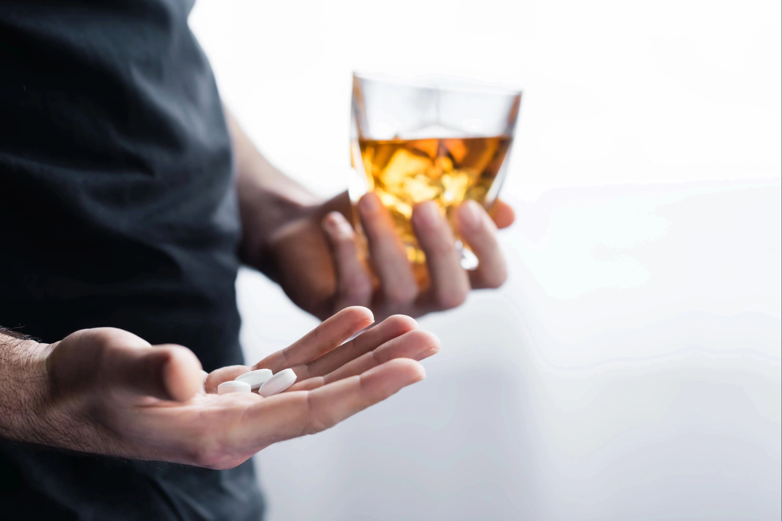Person holding pills in one hand and a glass of amber liquid in the other.