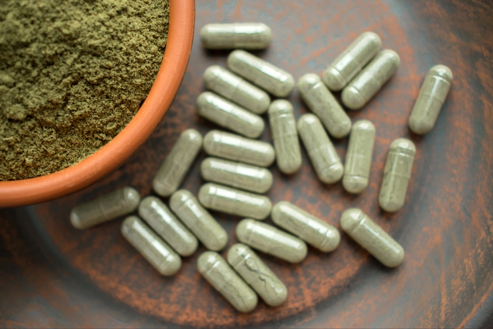 Kratom Withdrawal: Bowl of green powder next to scattered green capsules on a brown surface.